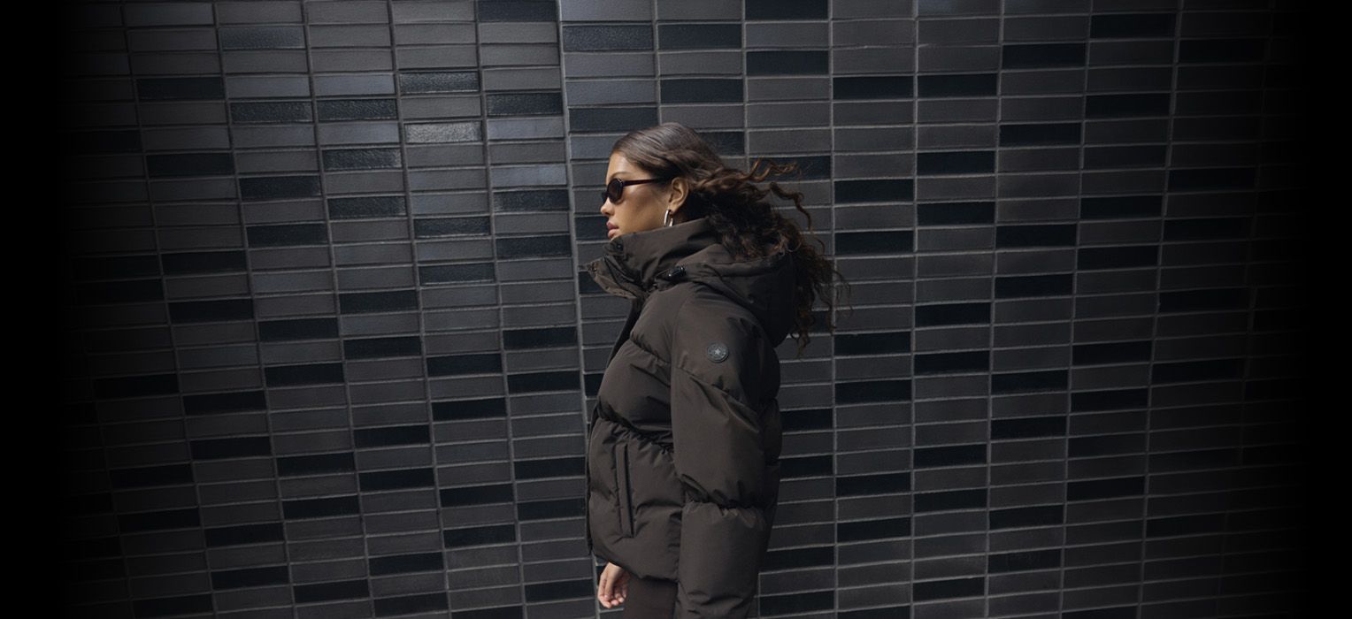 Person in a black puffer jacket and sunglasses walking against a dark tiled wall.