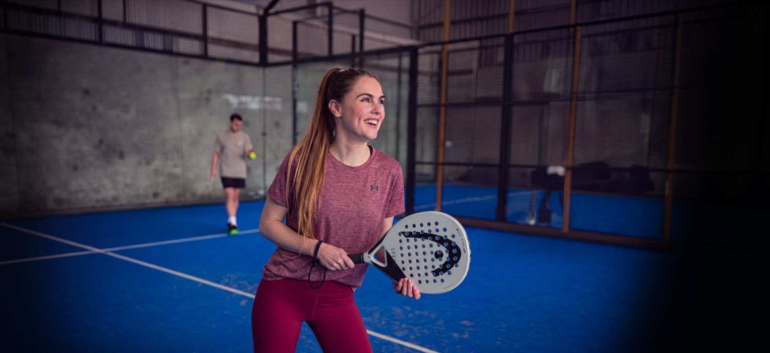 A woman in athletic wear smiles while holding a padel racket on an indoor court, with a blurred player in the background.