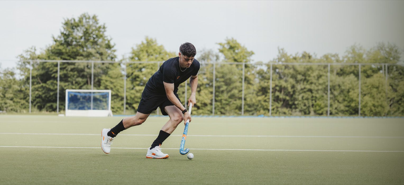 A man in black sports attire playing field hockey on a green turf, focused on hitting the ball with a blue hockey stick.