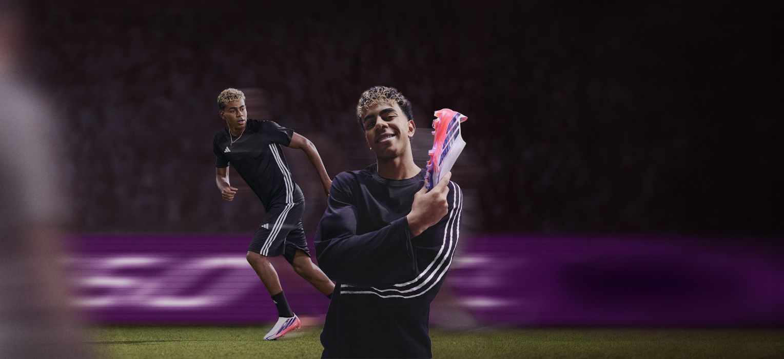 Soccer player wearing a black outfit, running on the field and holding a colorful cleat, smiling, with a stadium backdrop.