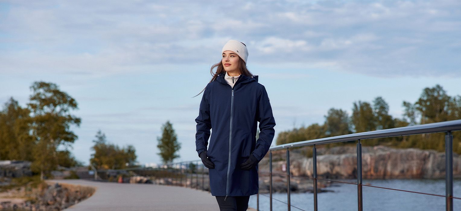 A woman in a navy coat and white beanie walks along a waterfront path with trees and rocks in the background under a cloudy sky.