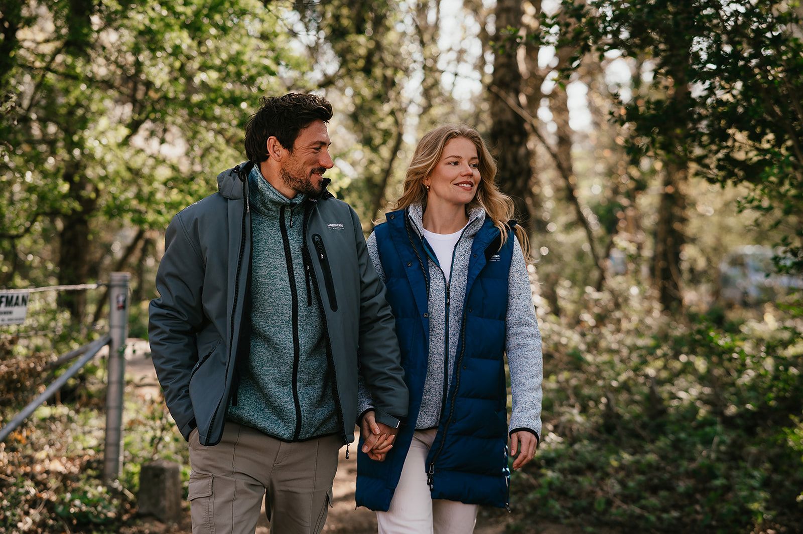 A couple walks hand in hand through a lush, sunlit forest, both wearing warm Nordberg outdoor jackets and smiling contentedly.