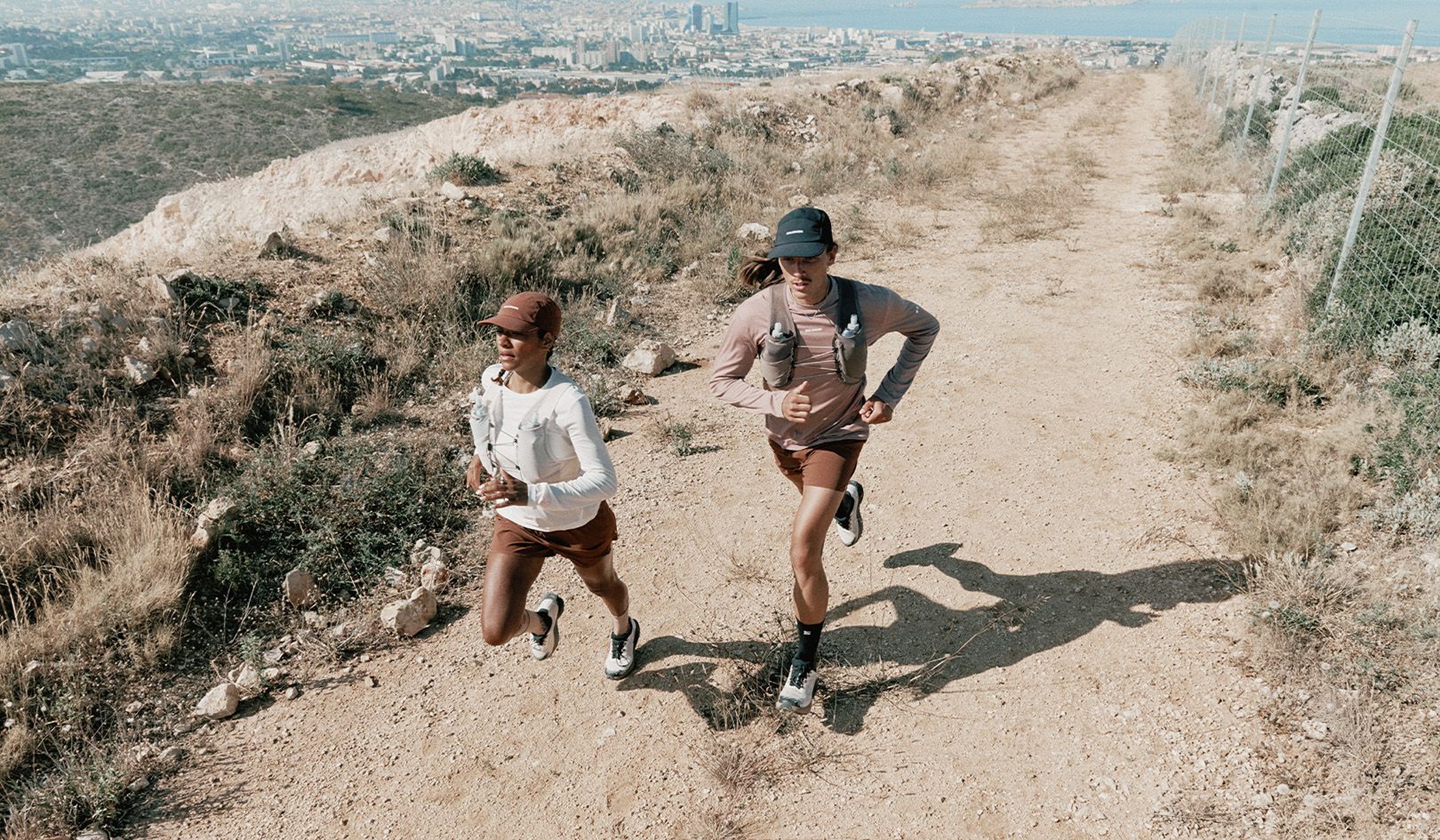 Two people in athletic gear trail running on a dirt path, with a cityscape and ocean in the background.