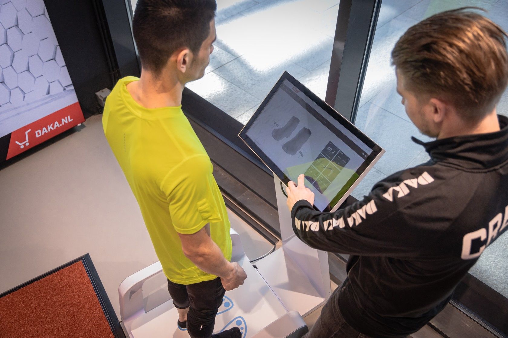 A person in a bright green shirt stands on a foot scanning device while another person operates a touchscreen displaying foot images.