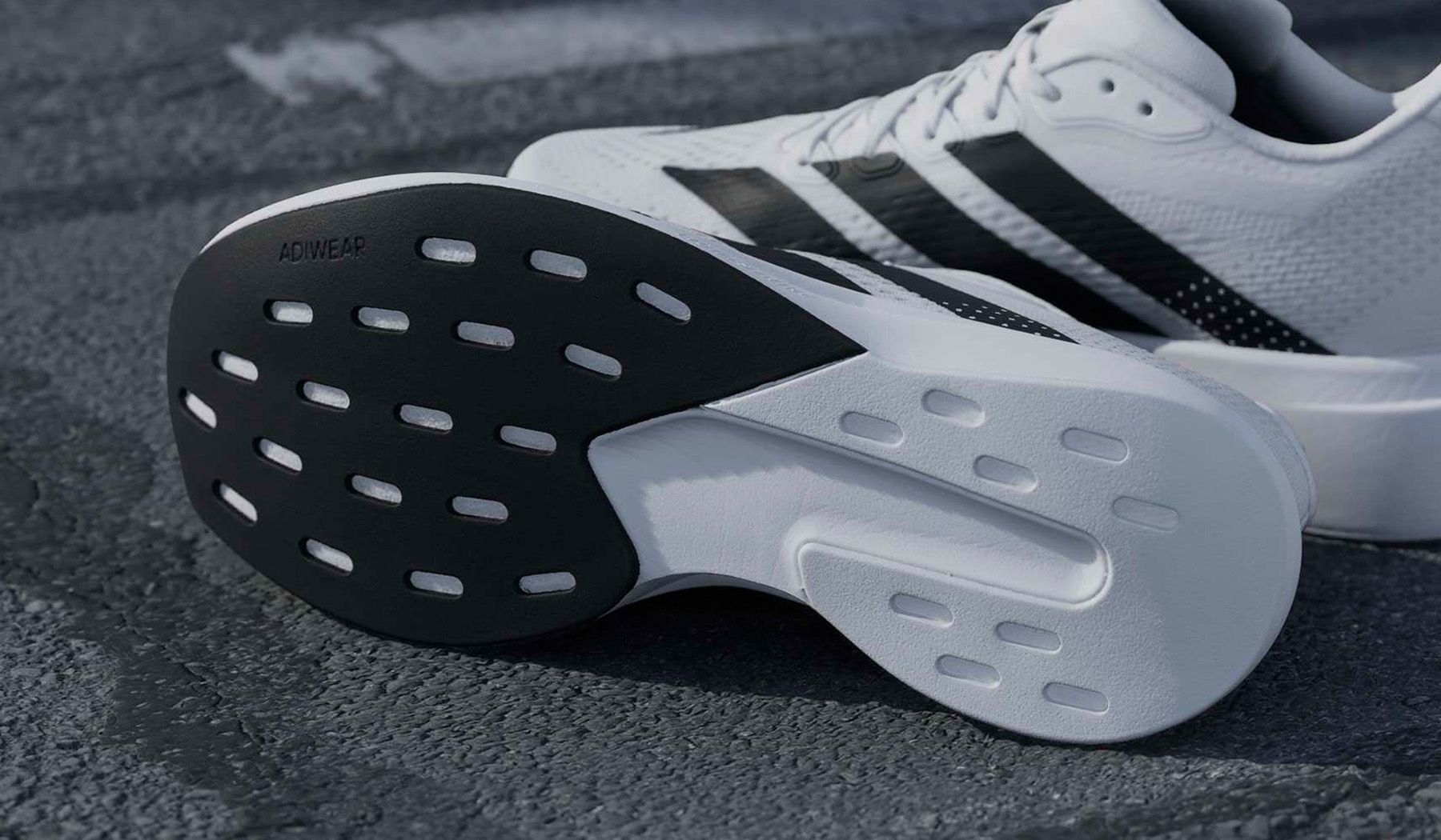 Close-up of white sneakers with black stripes, focusing on the textured soles marked "ADIWEAR" on a rough gray surface.