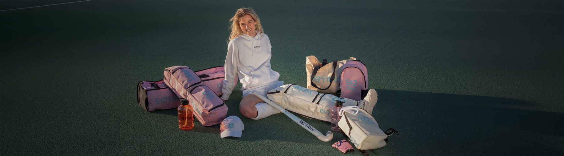 A person in sportswear sits on the ground surrounded by pink and white sporting bags and accessories.