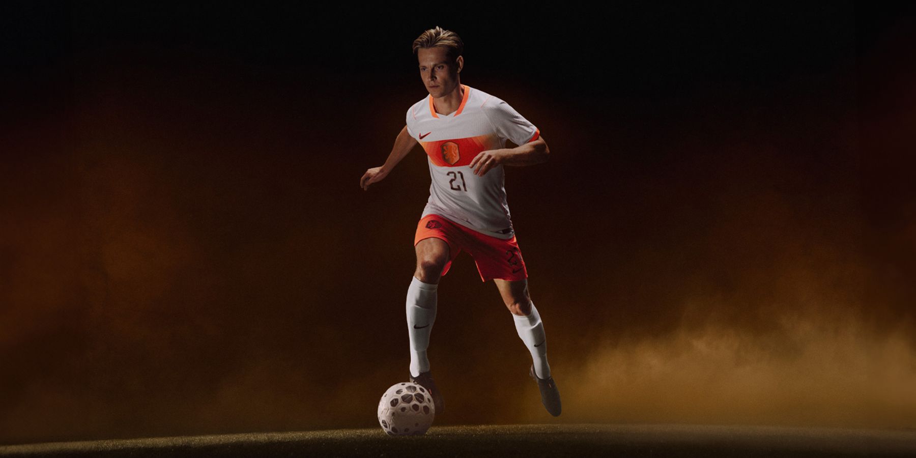 Soccer player in a white and orange uniform with number 21, dribbling a ball on a dark, smoky background.