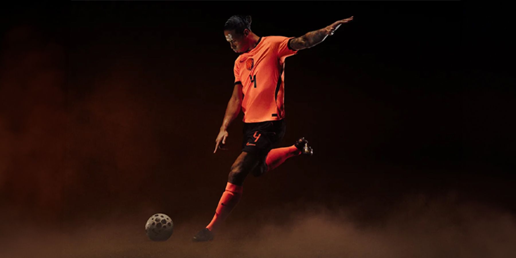 Soccer player in an orange jersey preparing to kick a ball, set against a dark, smoky background.