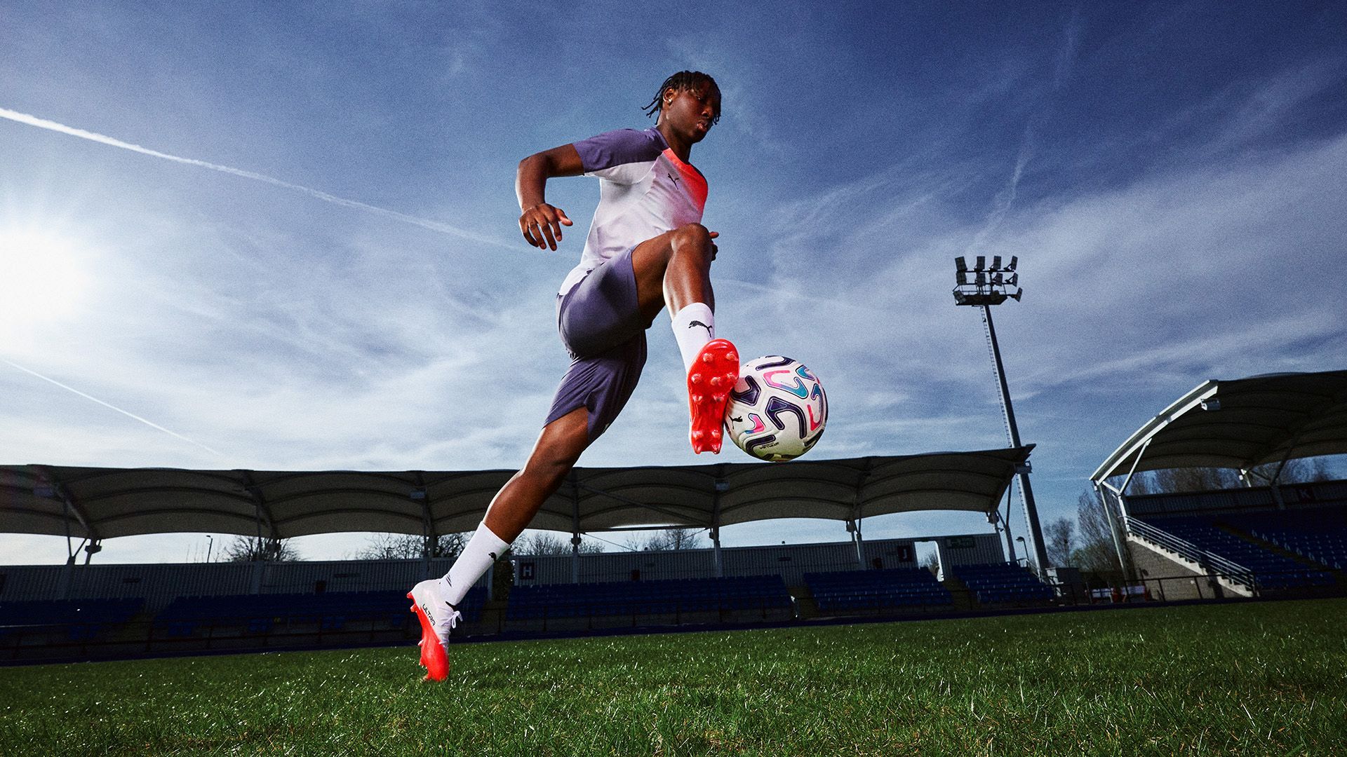 A soccer player leaps in the air, controlling a ball with his foot on a grassy field under a clear blue sky, with stadium seating in the background.