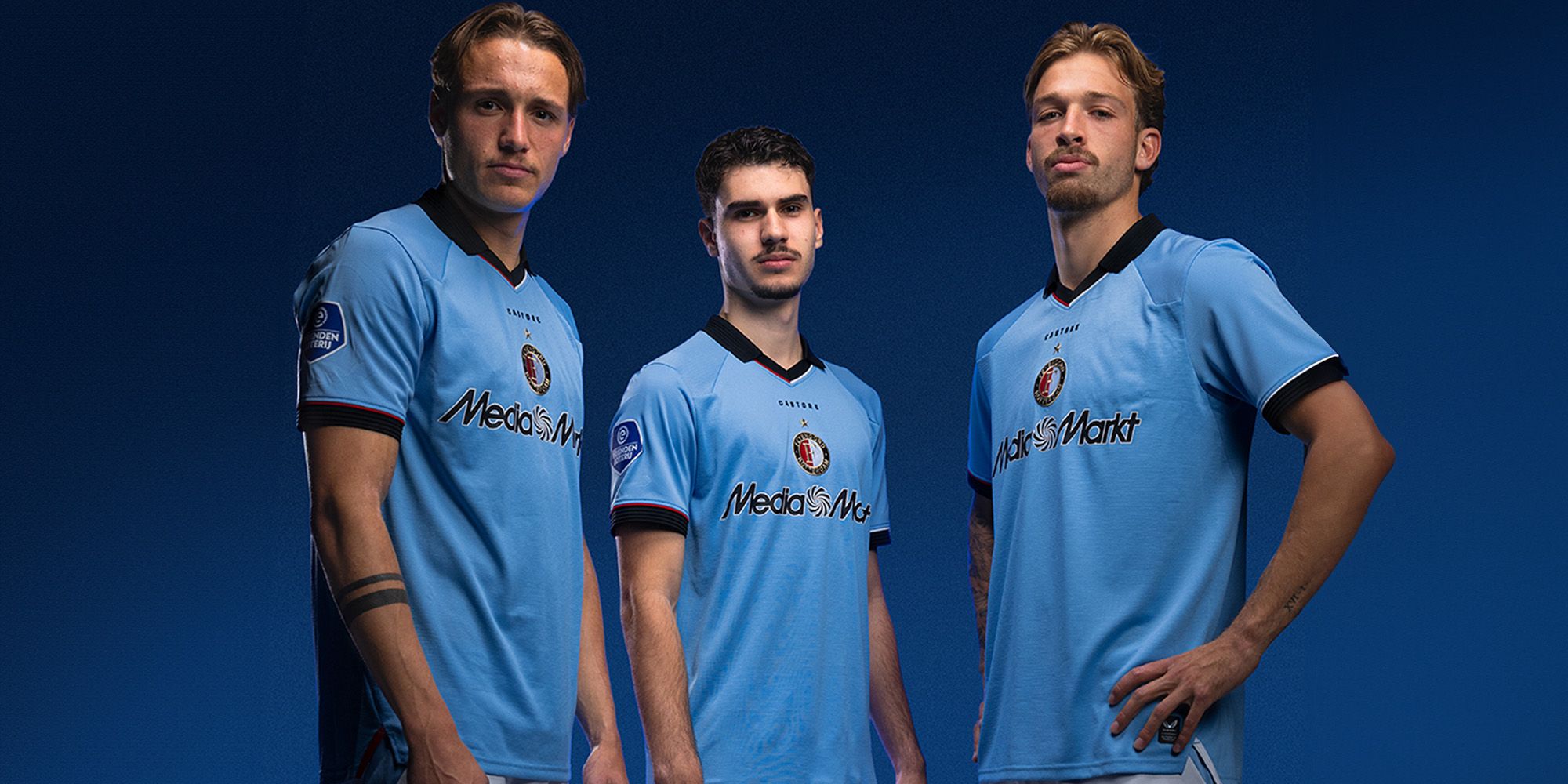Three soccer players in light blue jerseys pose confidently against a dark blue background.