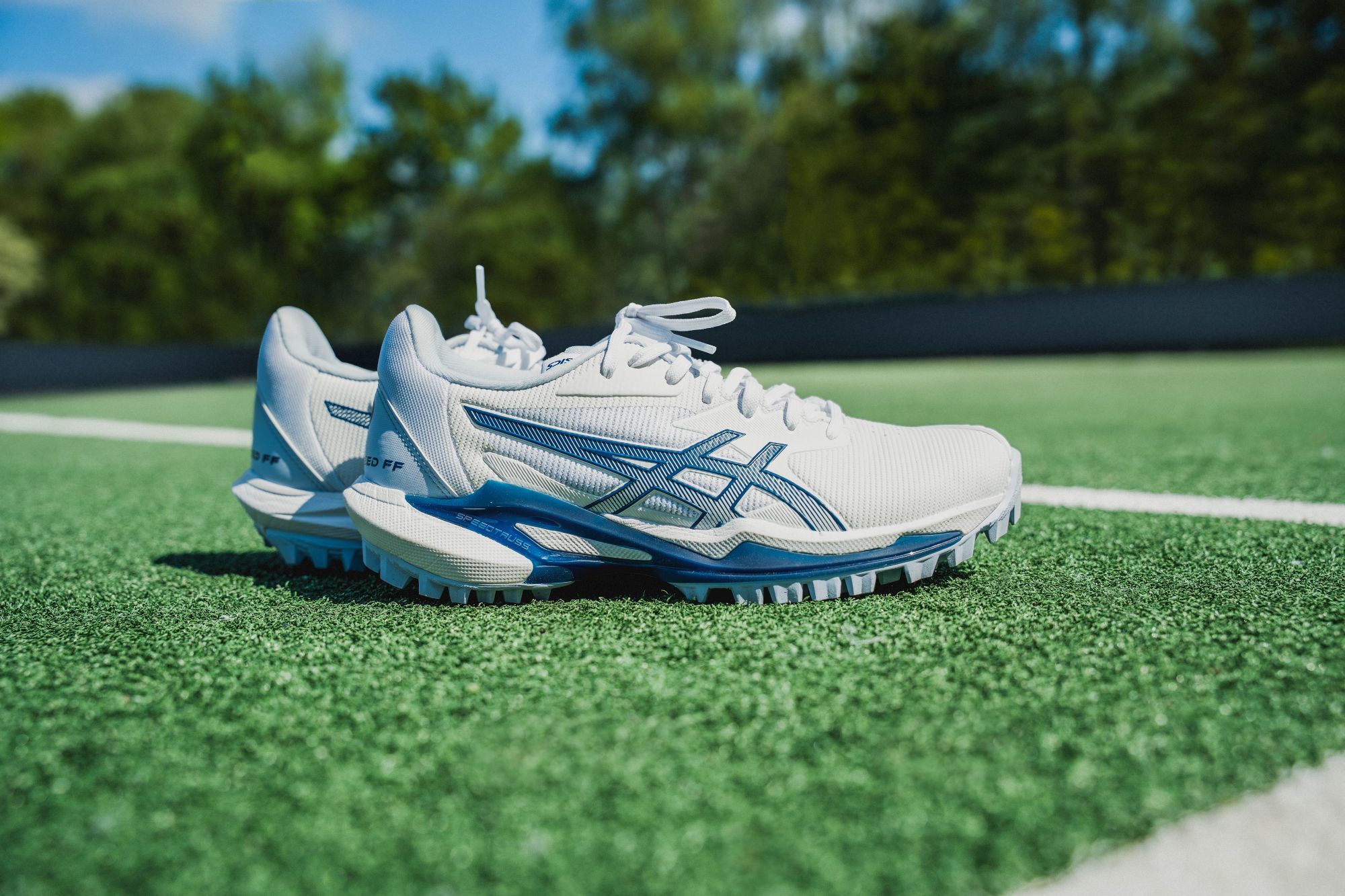 Hoe zorg je ervoor dat jouw hockeyspullen lang meegaan? White and blue Asics hockey shoes with textured soles on a green sports field, under a clear blue sky.