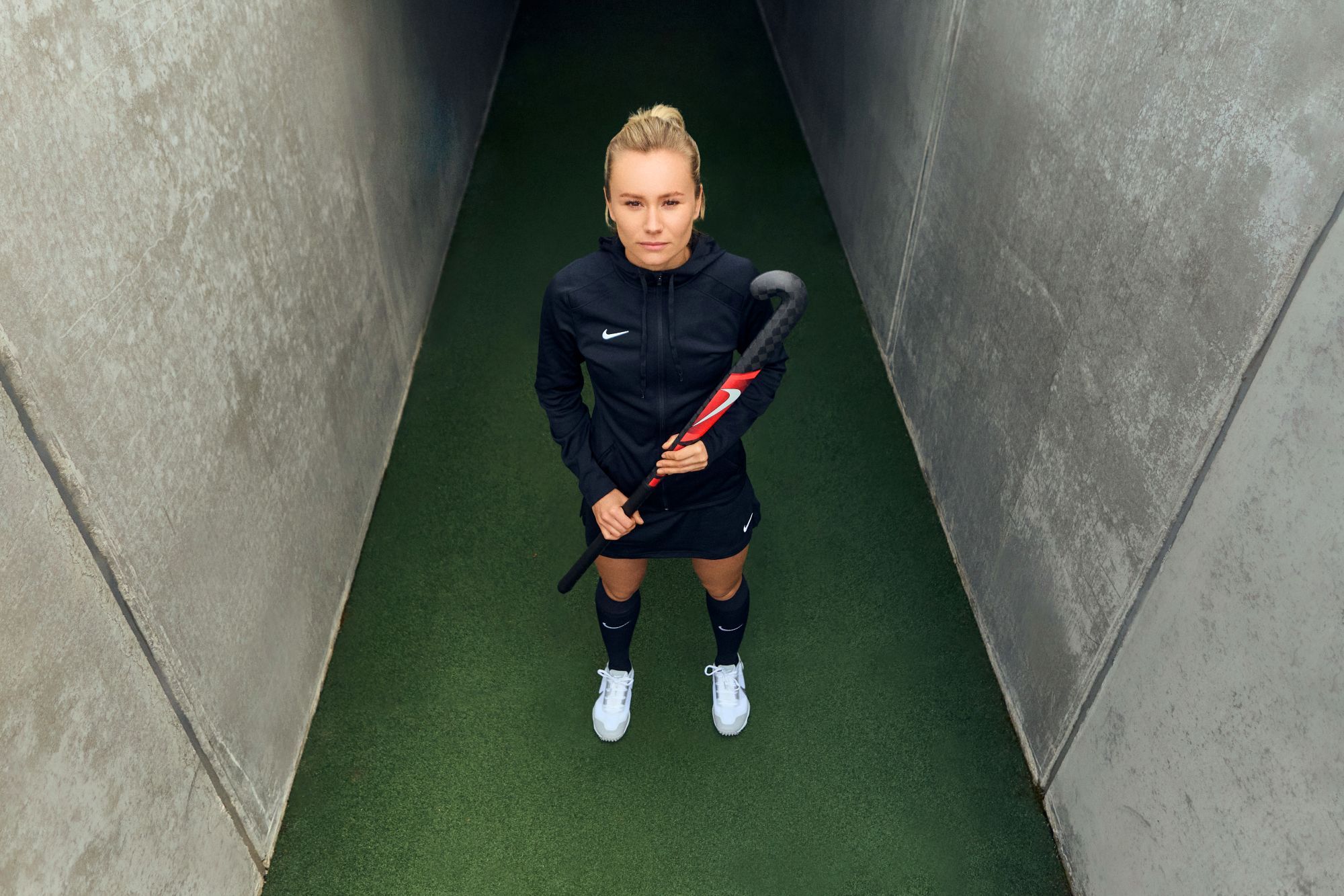 De evolutie van hockeysticks: hoe Nike de hockeywereld verandert A field hockey player stands confidently in a concrete tunnel, wearing a black Nike uniform and holding a hockey stick, viewed from above.