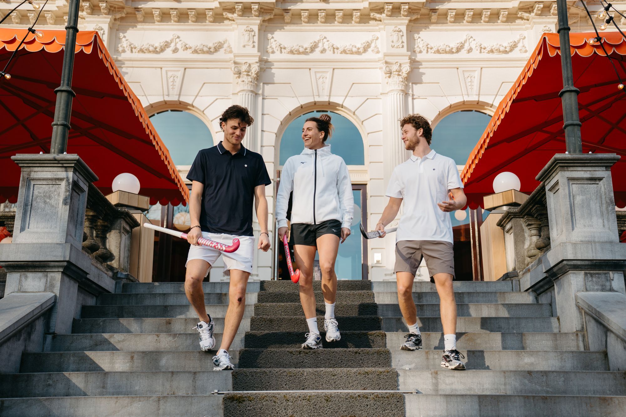 Welke hockeykleding heb je nodig? Three people holding Princess field hockey sticks walk down stone steps in front of a building with red awnings, smiling and chatting.