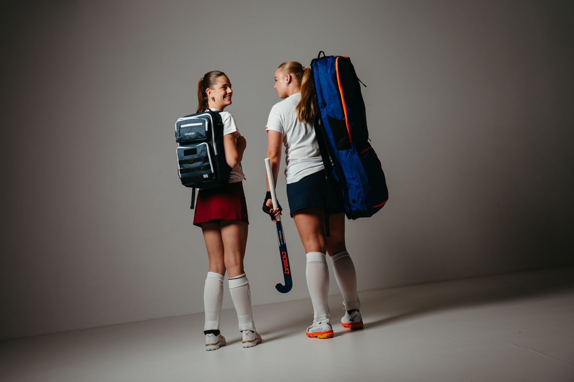 Hoe kies je de ideale sticktas voor hockey? Vind jouw perfecte tas Two field hockey players in uniform, holding Brabo equipment, look at each other, standing in a studio with backpacks.