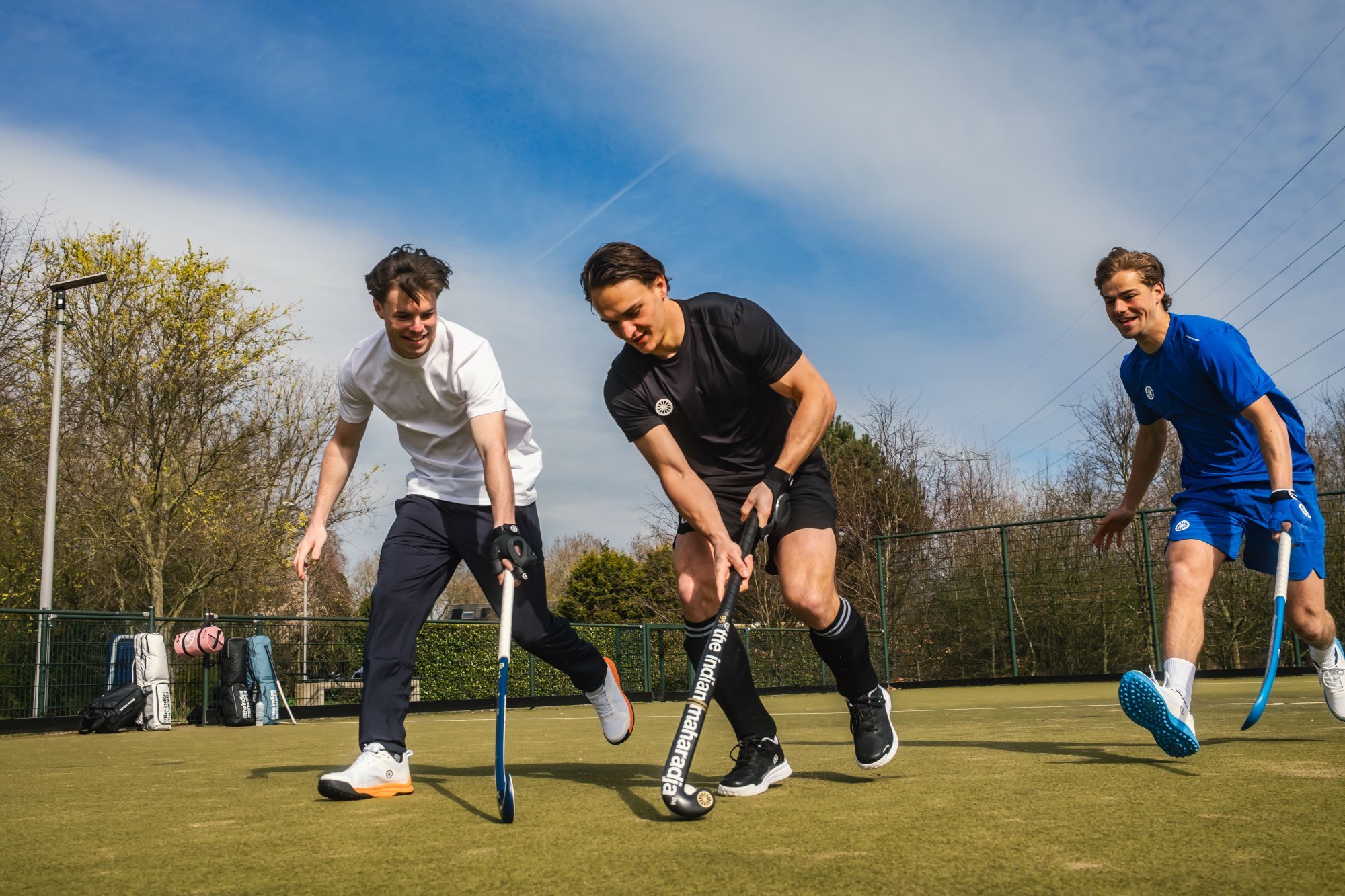 Hoe kies je de juiste kromming voor een hockeystick? Three men in sports attire playing field hockey on an outdoor pitch under a clear sky.
