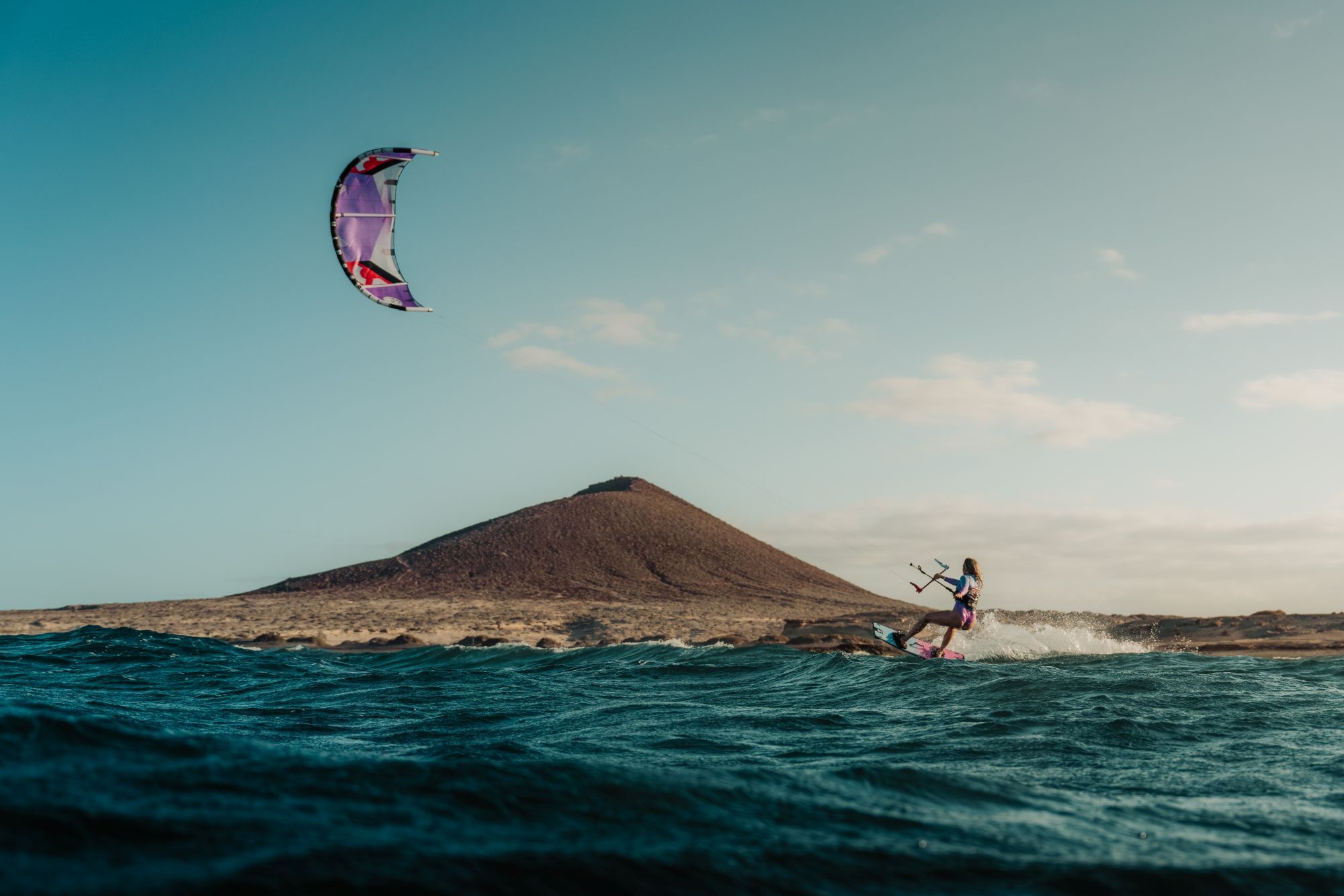 Kitesurfster snijdt door de golven van een donkerblauwe zee met een paarse vlieger en een vulkanische berg in de achtergrond.