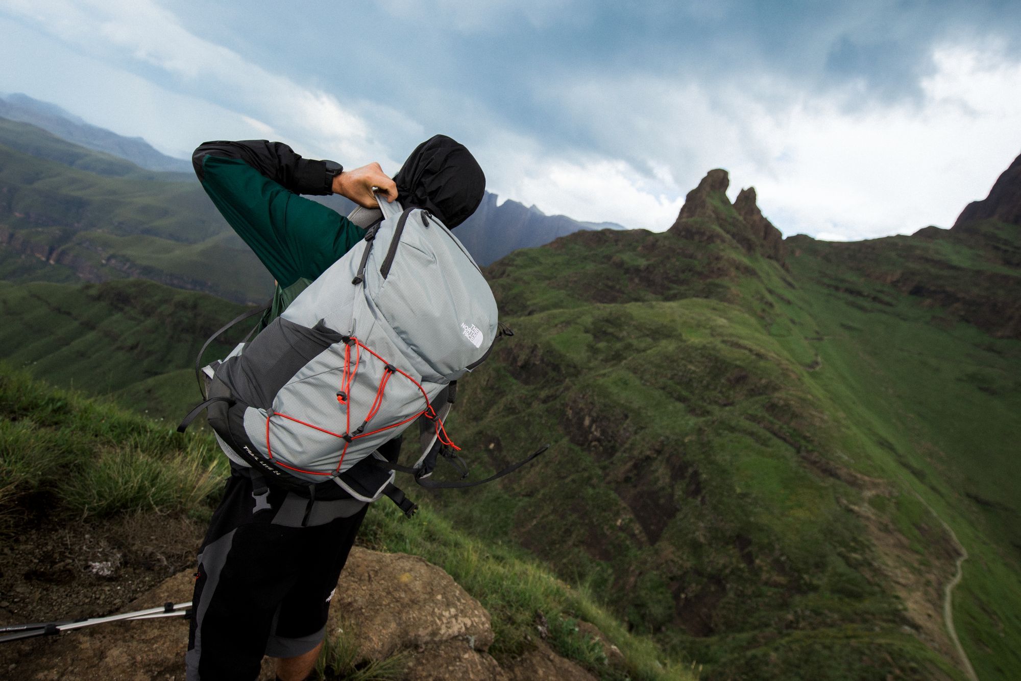 Wandelaar met een grijze The North Face Trail Lite rugzak kijkt uit over een uitgestrekt groen berglandschap.