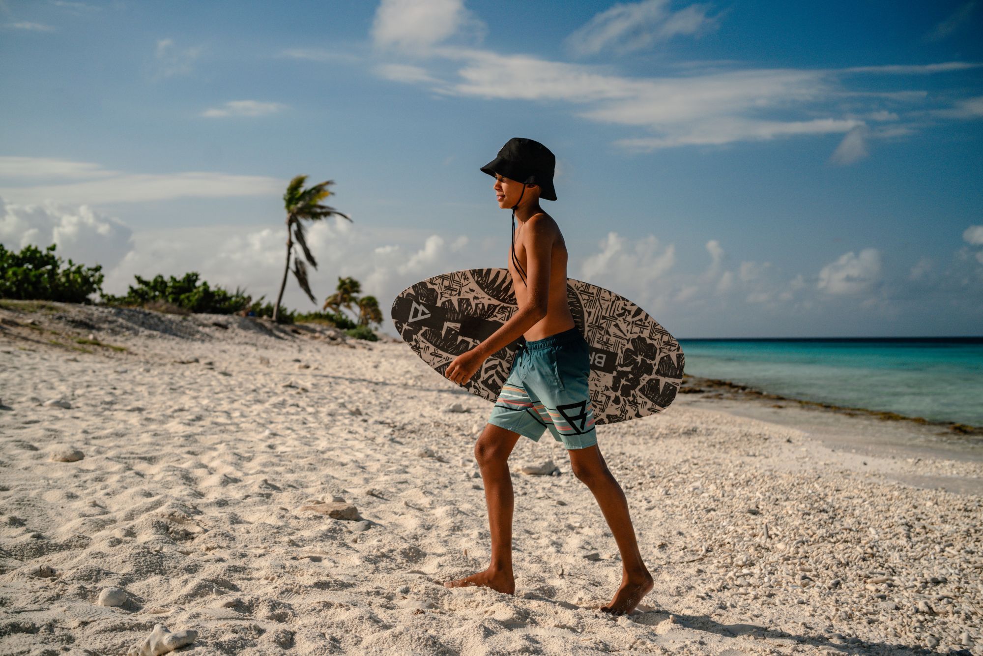 Jongen met een zwart vissershoedje loopt over een wit zandstrand met een bedrukt skimboard onder zijn arm langs een turquoise zee.