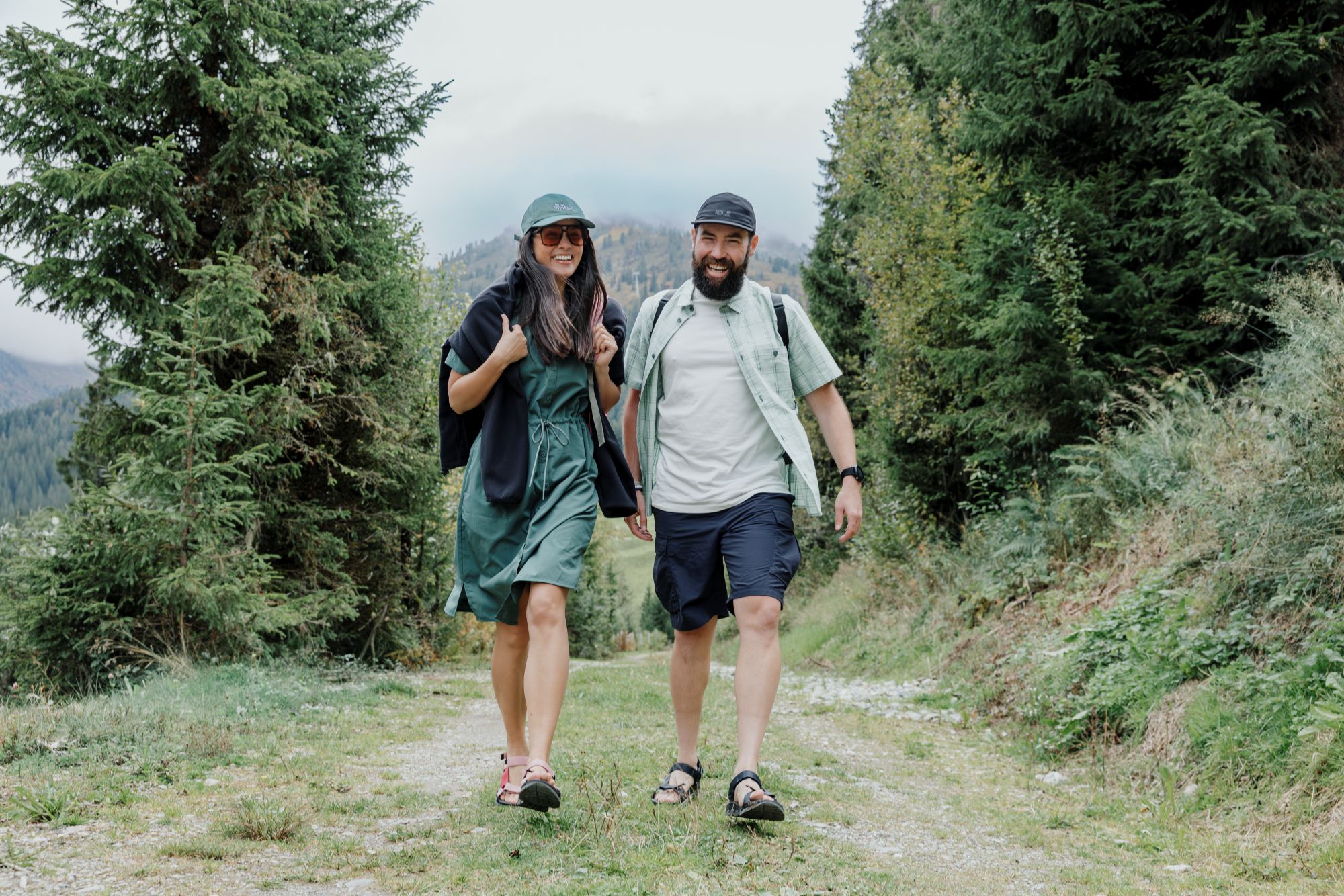 De complete paklijst voor je outdoor vakantie: Ga voorbereid op avontuur Lachende man en vrouw wandelen over een bospad in casual Jack Wolfskin zomerkleding en sandalen.