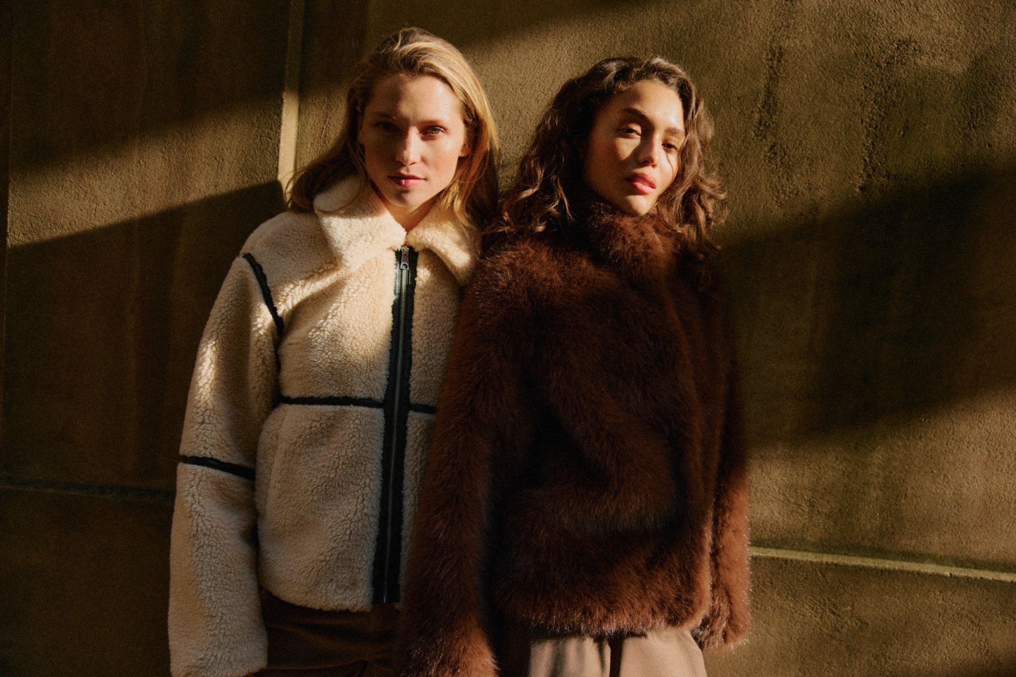 Two women wearing cozy fur Vero Moda jackets stand side by side against a warmly lit wall, casting shadows.