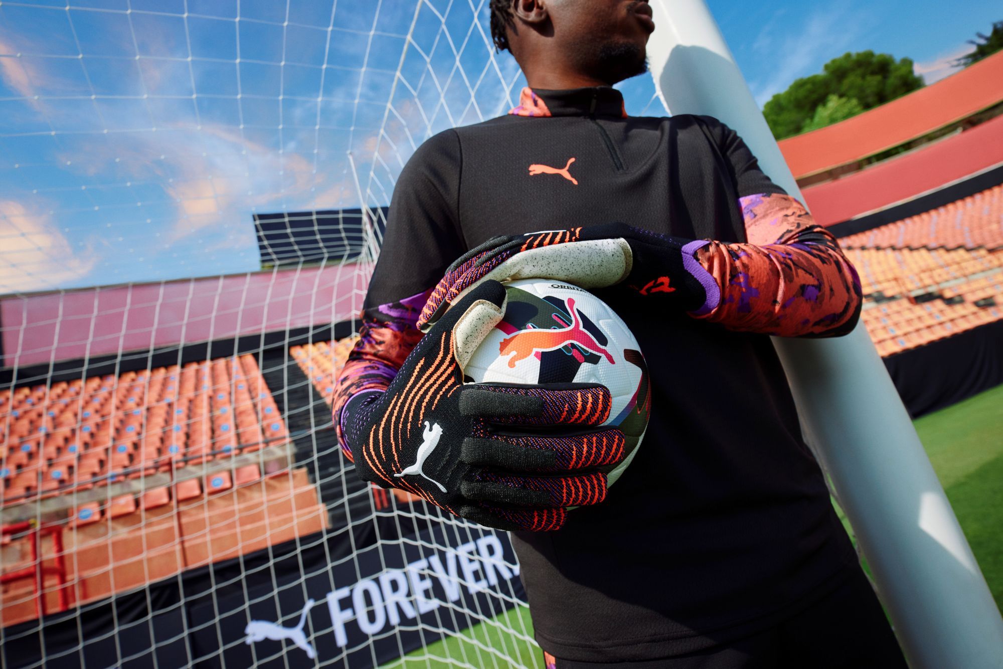 Soccer player in colorful Puma goalkeeper gloves holds a ball, standing near the goalpost in a stadium.