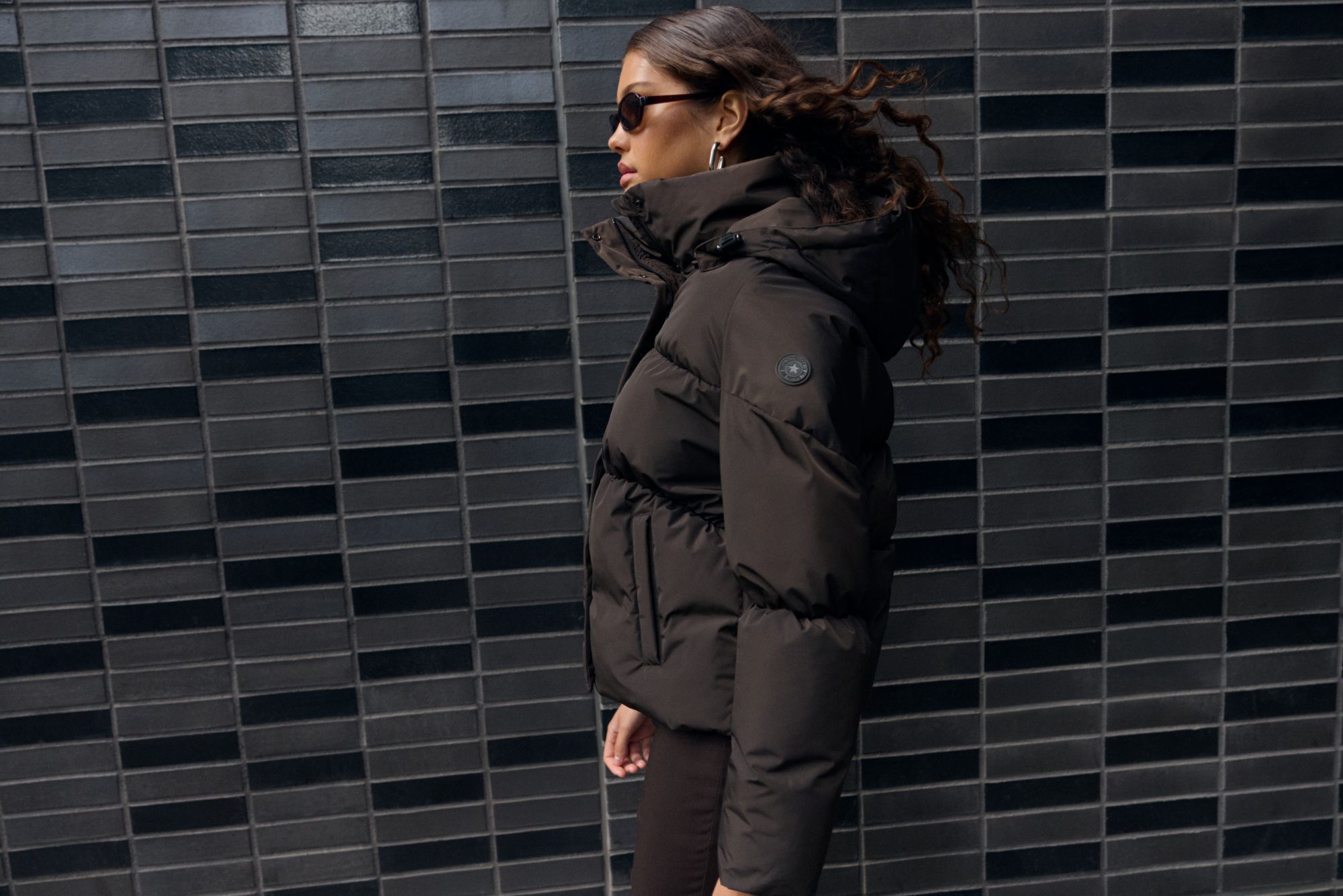 A woman wearing sunglasses and a brown Airforce puffer jacket walks past a tiled wall, with her hair blowing in the wind.