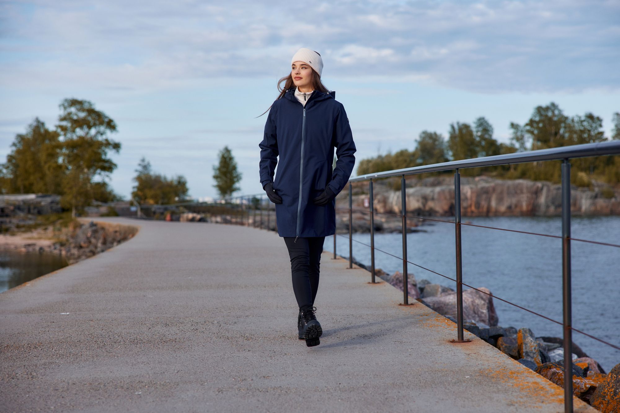 Winterjassen Trends Herfst/Winter 2026: Vind Jouw Perfecte Match A woman in a navy Luhta coat and white beanie walks along a scenic waterfront path with trees and rocks in the background.