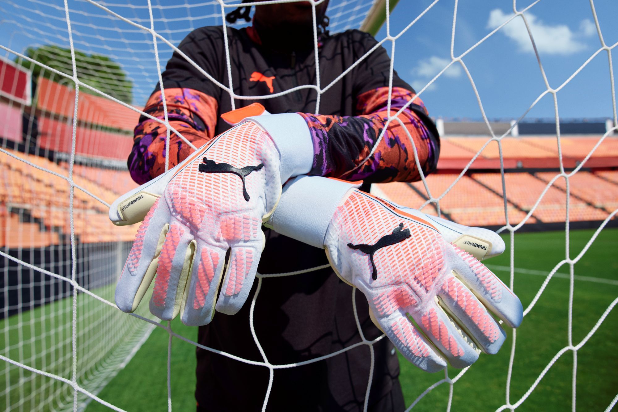 Hoe kies je de juiste keepershandschoenen? Vind jouw perfecte match A person wearing orange and purple Puma goalkeeper gloves crosses hands in front of a soccer goal net in an empty stadium.