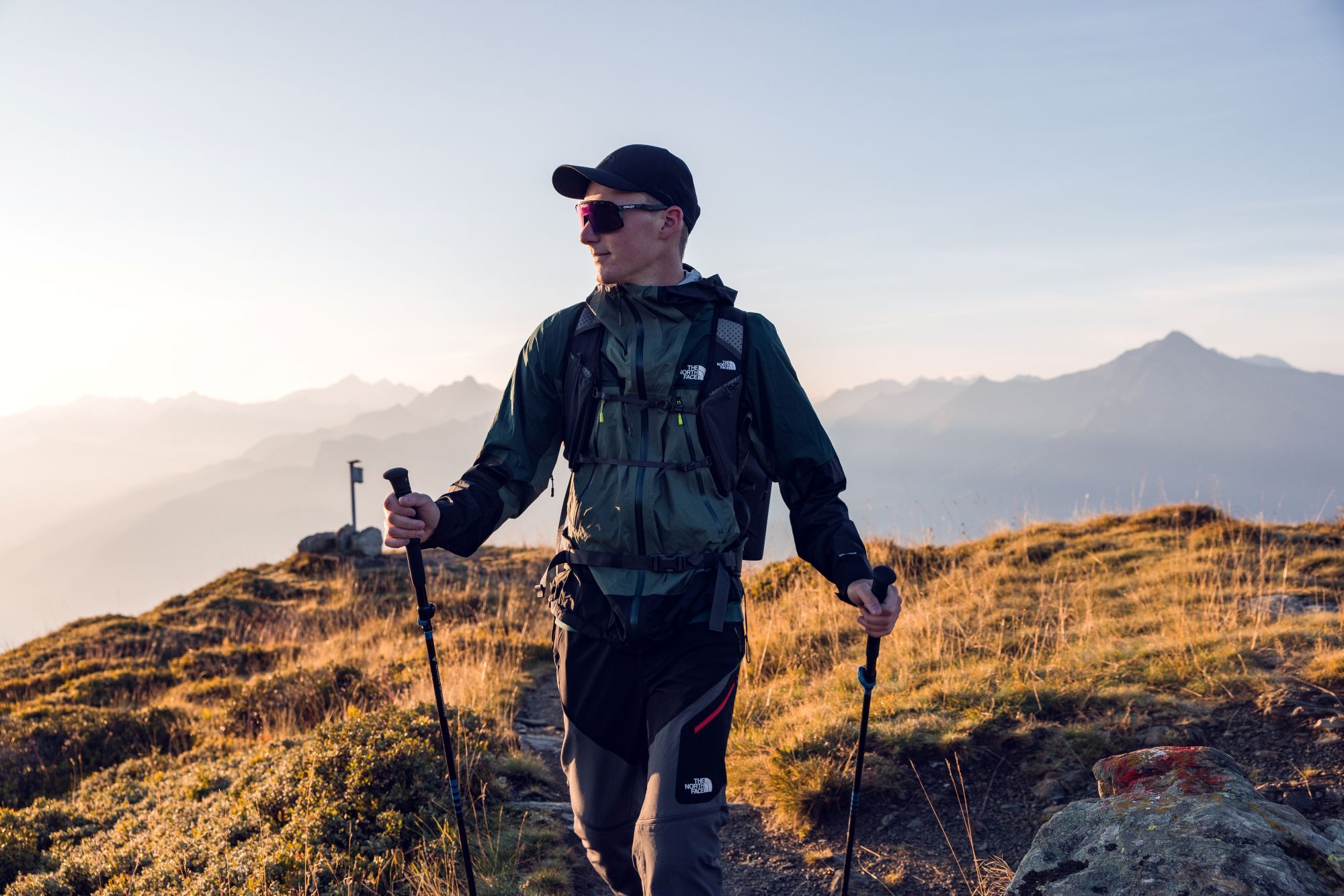 Man in The North Face jas met wandelstokken en zonnebril loopt op een bergkam tijdens zonsondergang.
