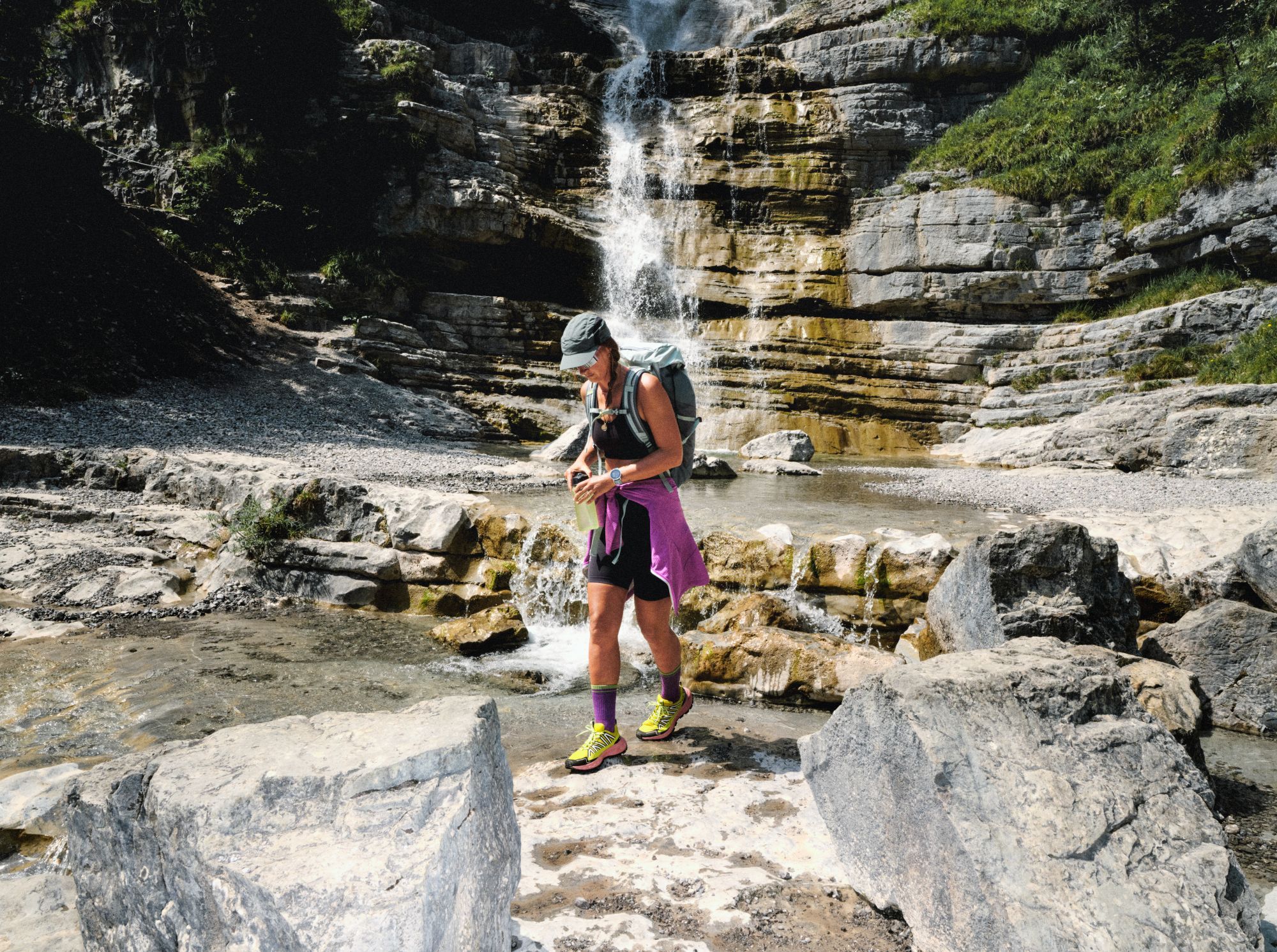 Vrouw in Jack Wolfskin sportkleding met rugzak wandelt langs een waterval over rotsachtig terrein.