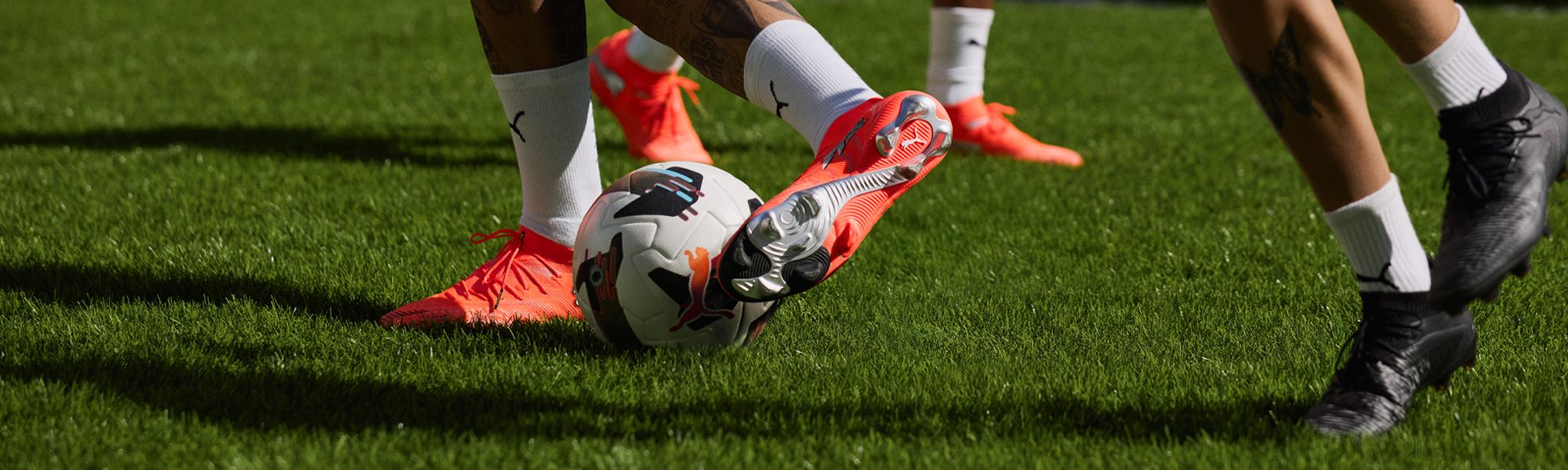 Close-up of soccer players competing for the ball on a grassy field, wearing bright orange and black cleats.