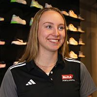 Smiling woman with long blond hair wearing a black polo shirt in a shoe store, with various sneakers displayed on the shelves behind her.
