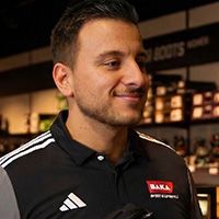 Man in a black and gray sports shirt stands in a store, smiling. Shelves with various products are visible in the background.