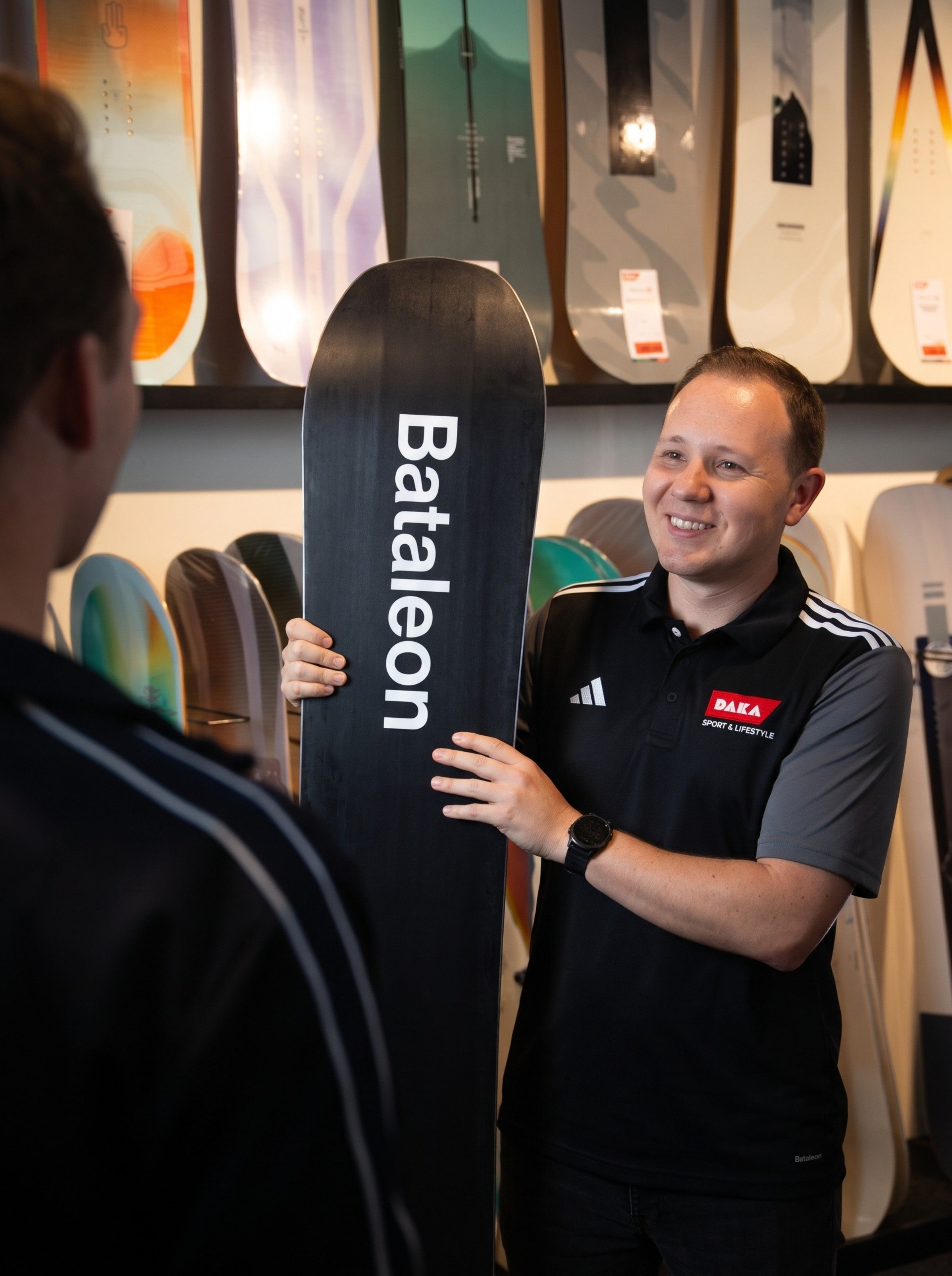 A person holding a snowboard with the brand name "Bataleon" in a shop, smiling, surrounded by various snowboard designs.