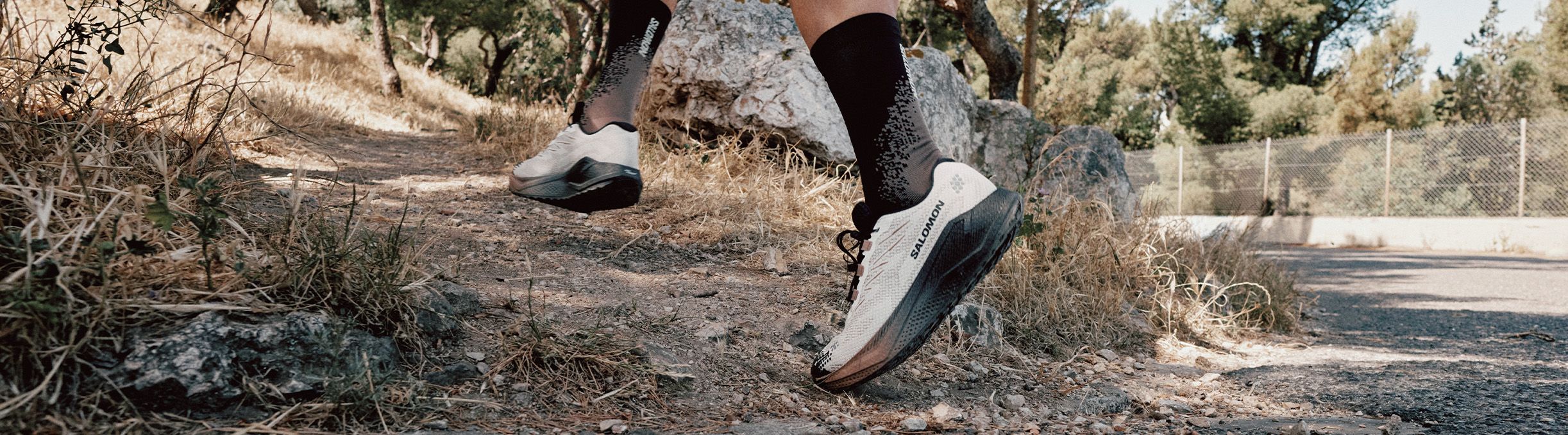 Close-up of a person in white running shoes and black socks on a rocky trail, with trees and grassy terrain in the background.