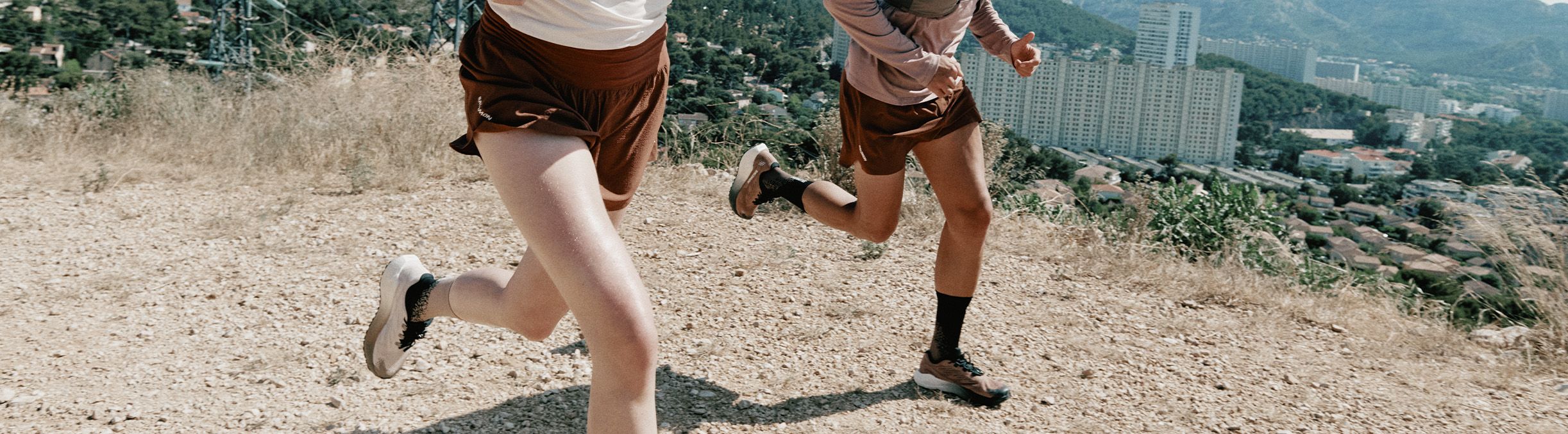 Two people running on a gravel path with a cityscape and mountains in the background. They are wearing athletic clothing and sneakers.