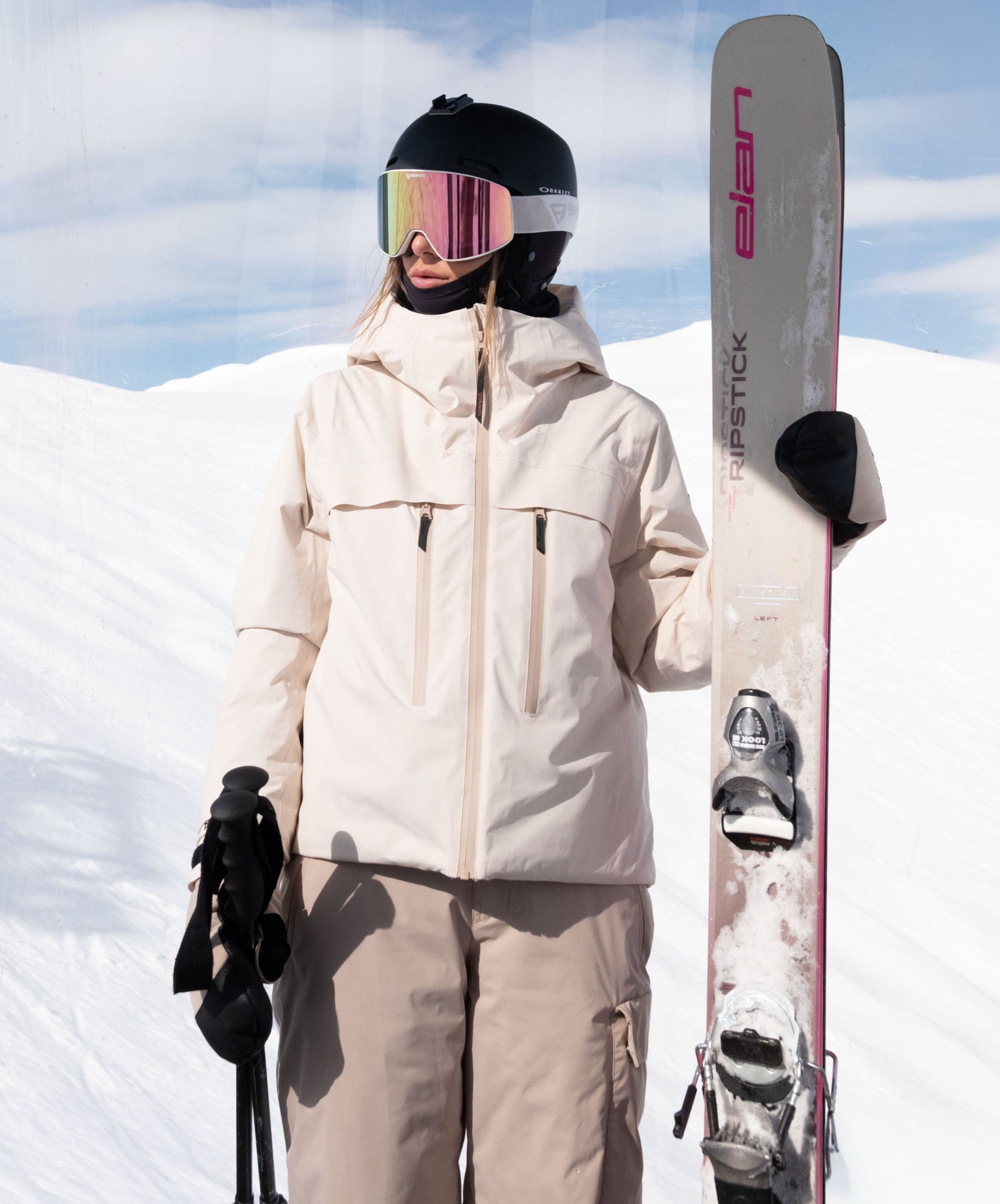 Zo Blijf jij Warm op de Piste | Uitleg 3-lagensysteem Person in winter gear holding skis on a snowy mountain, wearing a helmet and reflective goggles, under a partly cloudy sky.