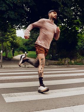 A bearded man in a cap and athletic wear runs across a crosswalk, surrounded by lush greenery.