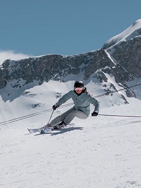 A skier in a gray jacket and helmet glides down a snowy slope with mountains in the background under a clear blue sky.