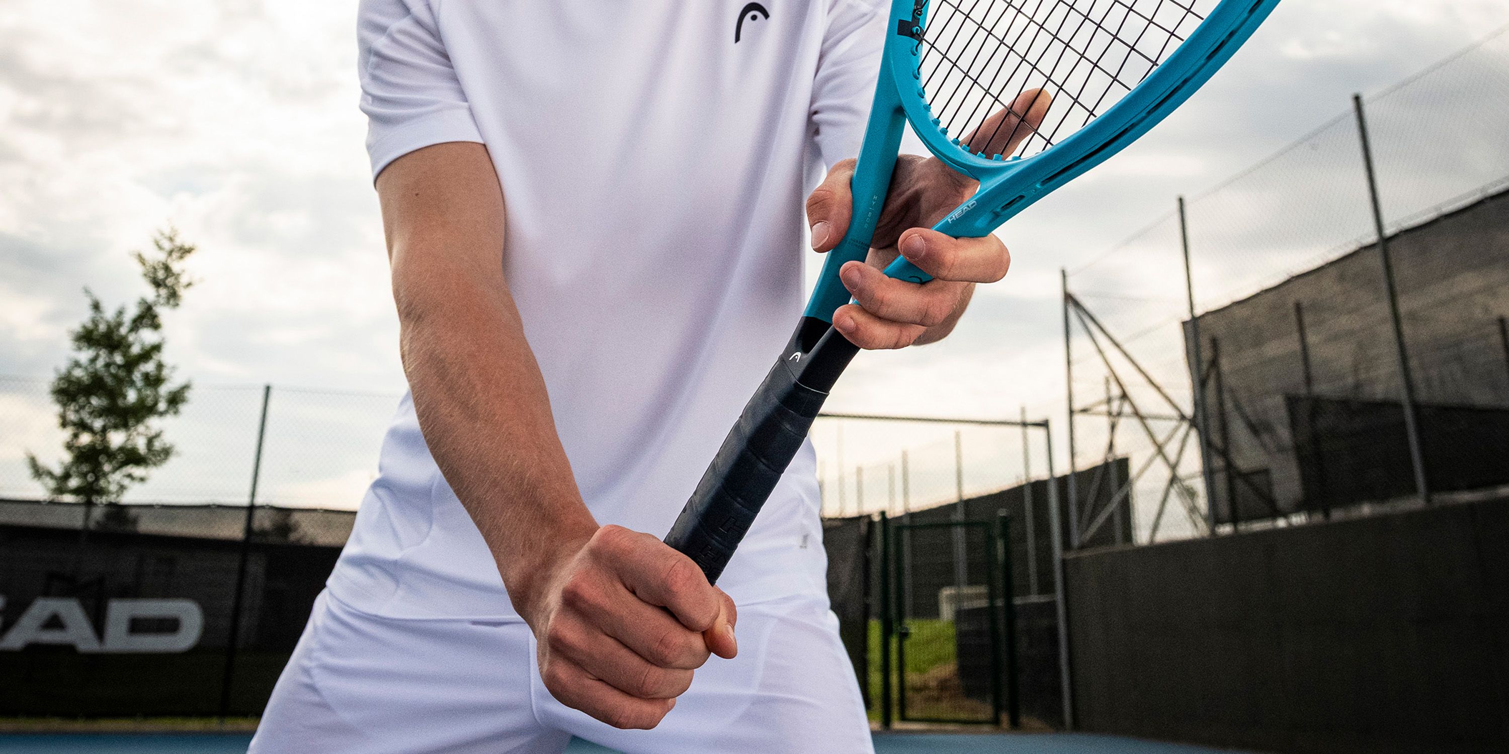 Close-up of a person in a white tennis outfit holding a blue tennis racket, preparing to serve on an outdoor court.