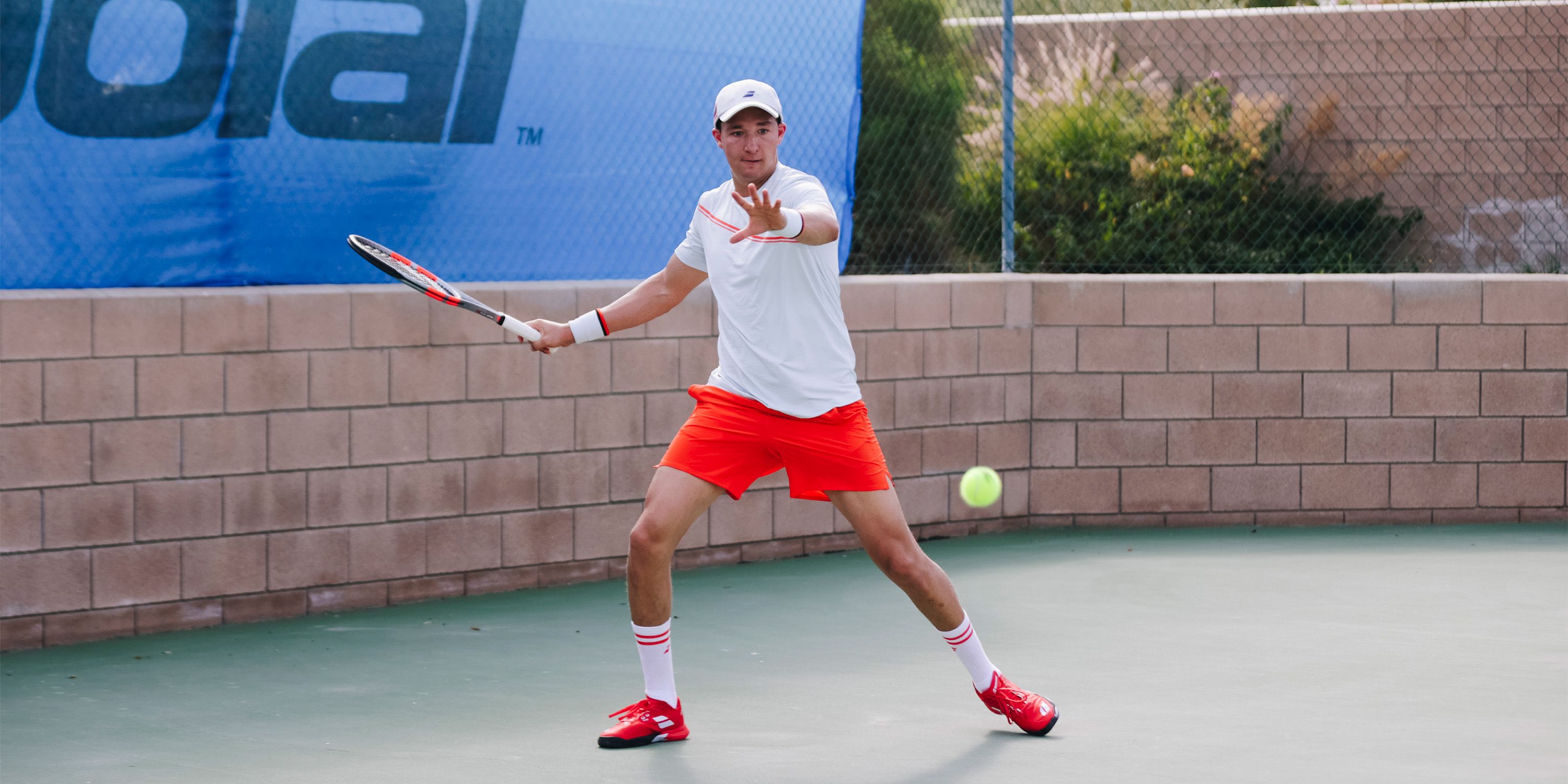 A tennis player in a white shirt and red shorts prepares to hit a ball with a racket on an outdoor court.