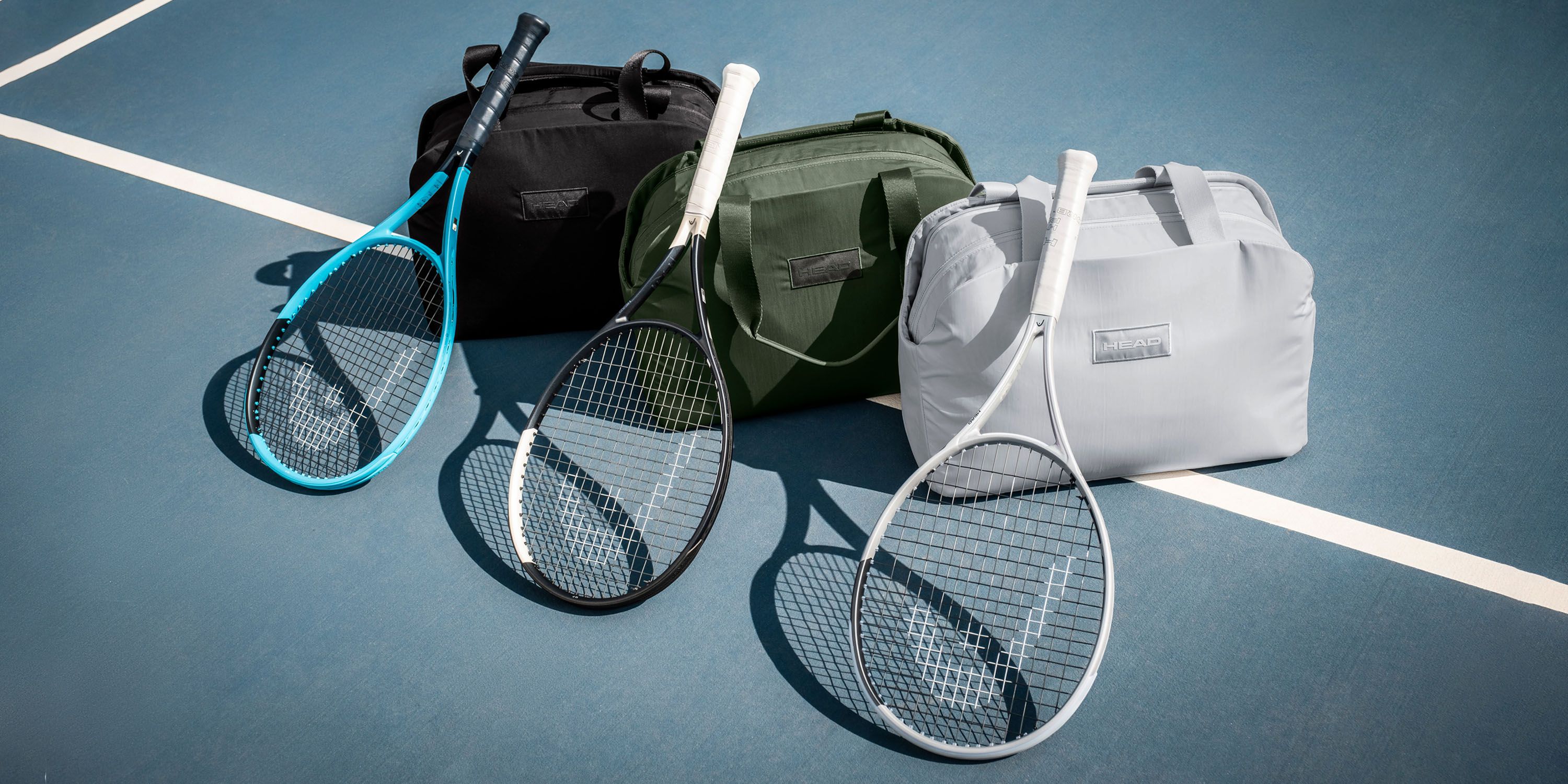 Three tennis rackets with different colored handles rest on a court surface, each paired with a matching bag in black, green, and gray.
