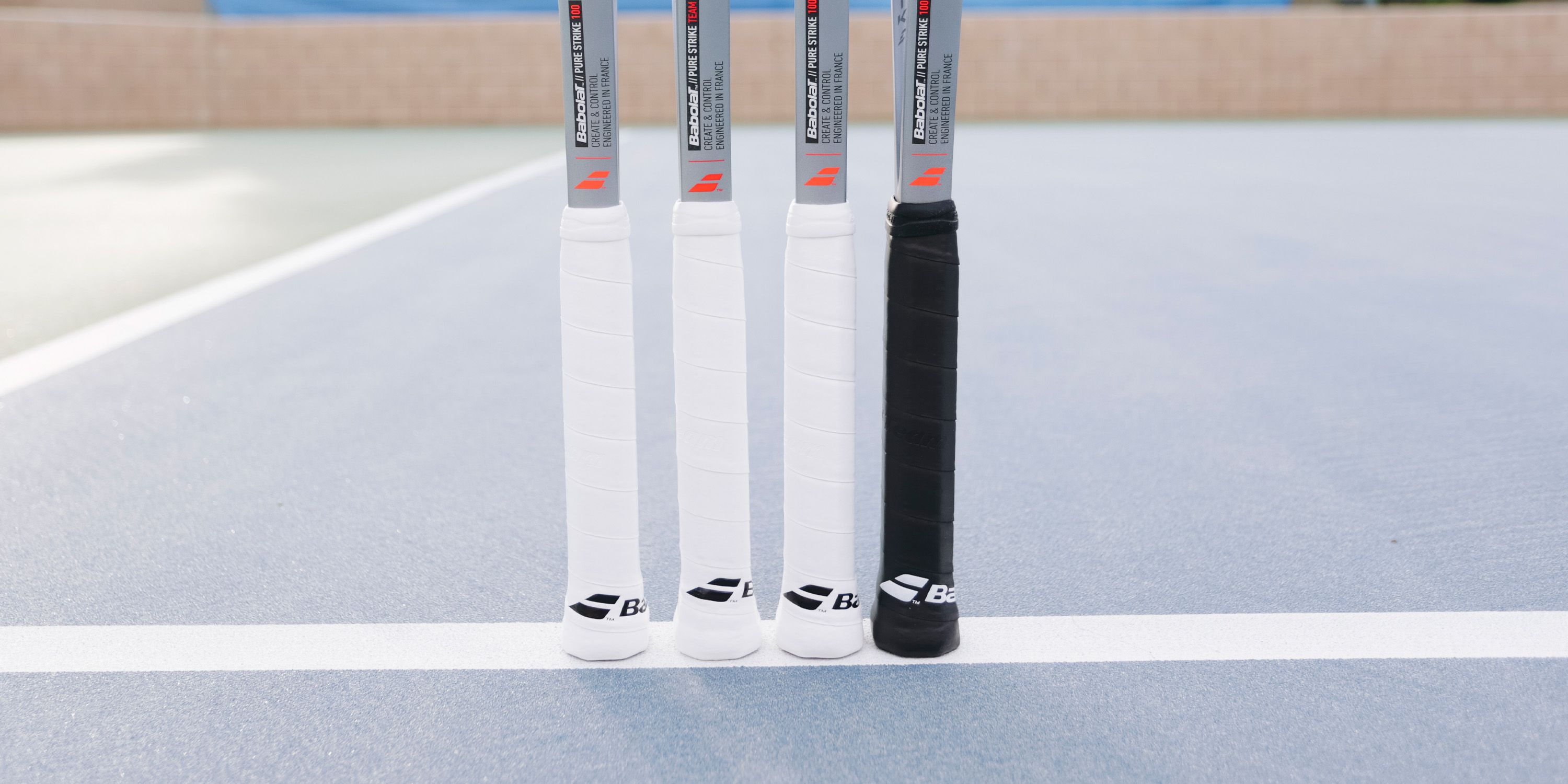 Four tennis racket handles on a blue court, three with white grips and one with a black grip, lying on a white line.