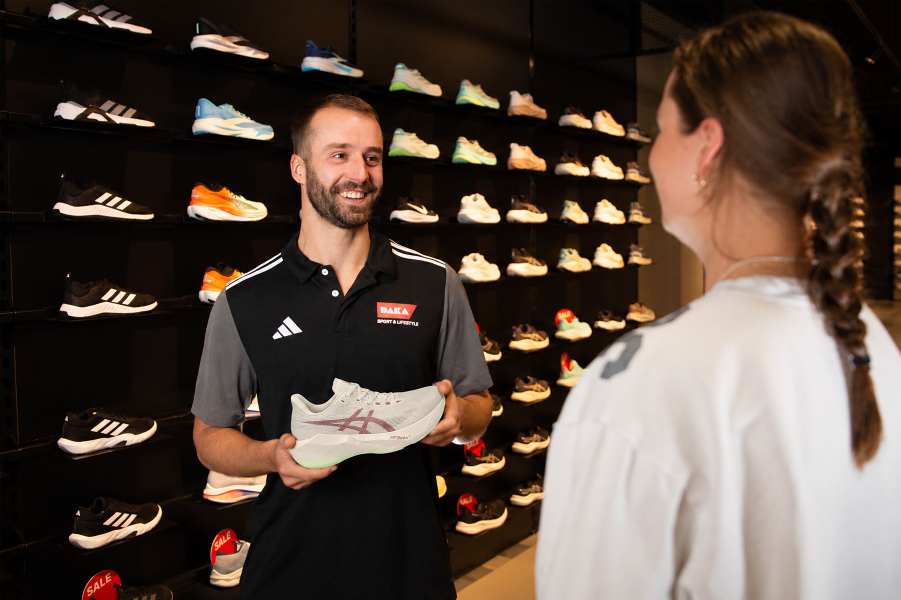 A salesperson holding a sneaker while talking to a customer in a shoe store with wall displays of various athletic shoes.