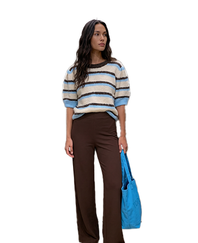 Dames Woman in striped sweater and brown pants holding a blue tote bag, standing against a plain background.