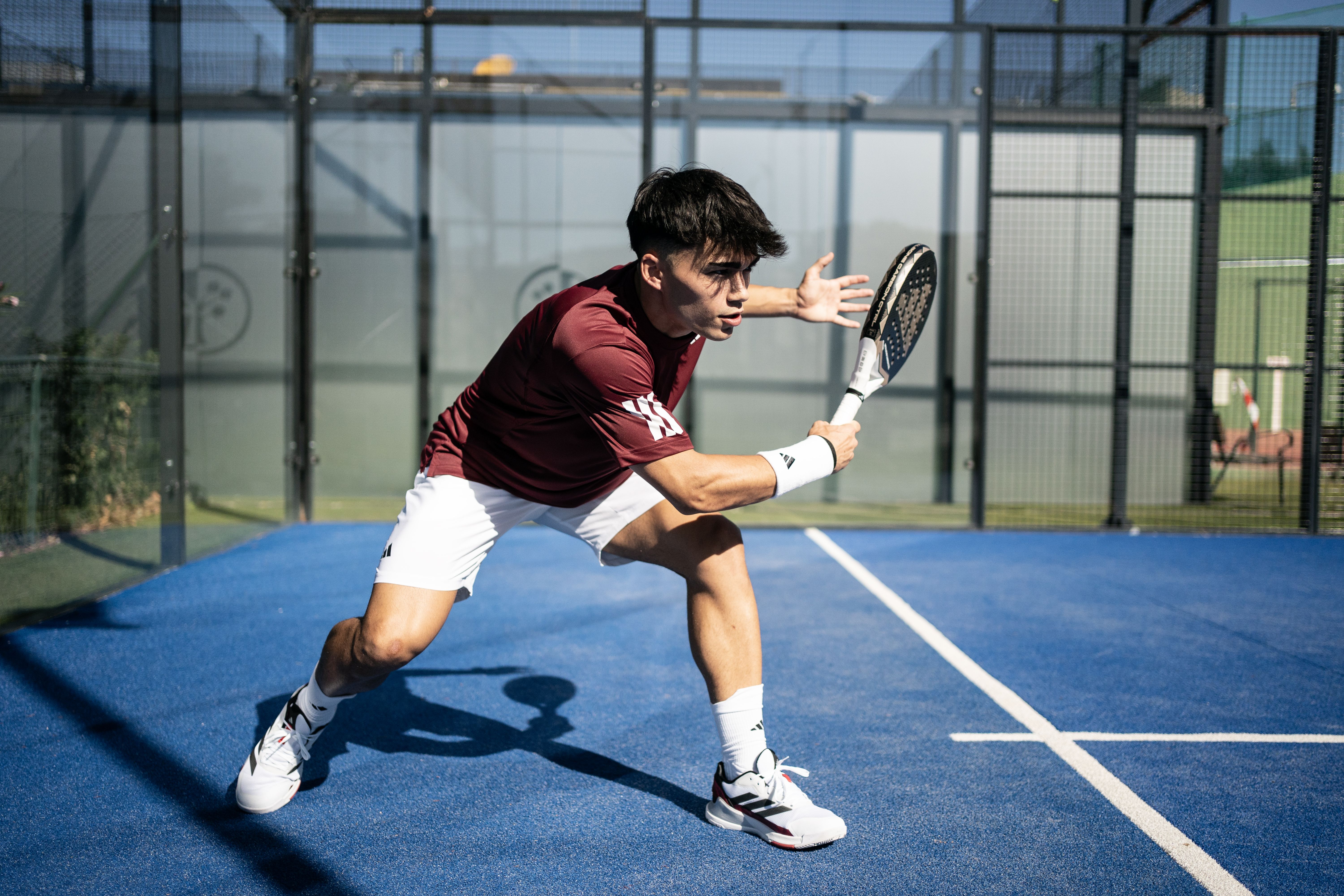 A tennis player in a maroon shirt and white shorts prepares to hit a shot on a blue court, with a focused expression and a ready stance.
