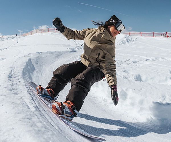 Person snowboarding down a snowy slope, wearing a beige jacket, black pants, and goggles, with a clear blue sky in the background.