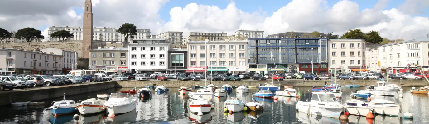 Le port de Brest animé avec de nombreux bateaux à quai, avec en toile de fond des bâtiments et une grande tour, sous un ciel partiellement nuageux.