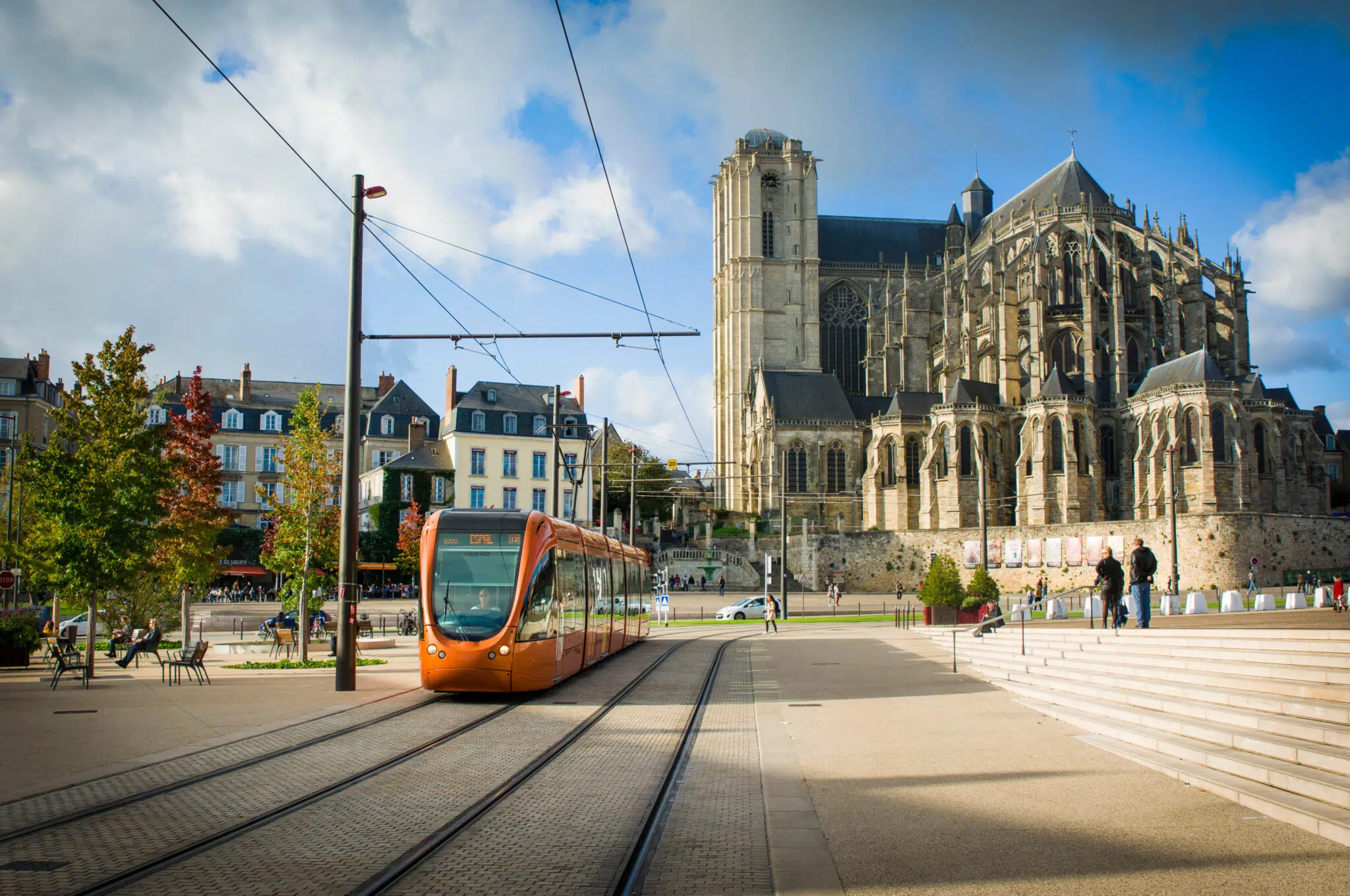 Tramway orange passant devant la cathédrale de la ville du Mans par une journée ensoleillée, avec des personnes marchant à proximité et des bâtiments colorés en arrière-plan.