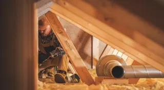 Personne travaillant dans des combles, entouré de poutres en bois et d'isolation, avec un conduit métallique visible.